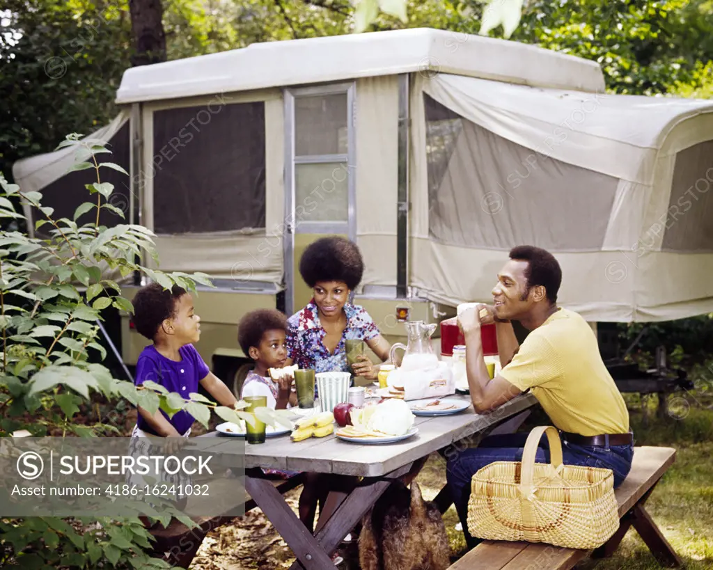 1970s SMILING AFRICAN-AMERICAN FAMILY EATING TOGETHER AT PICNIC TABLE IN FRONT OF POP-UP CAMPER