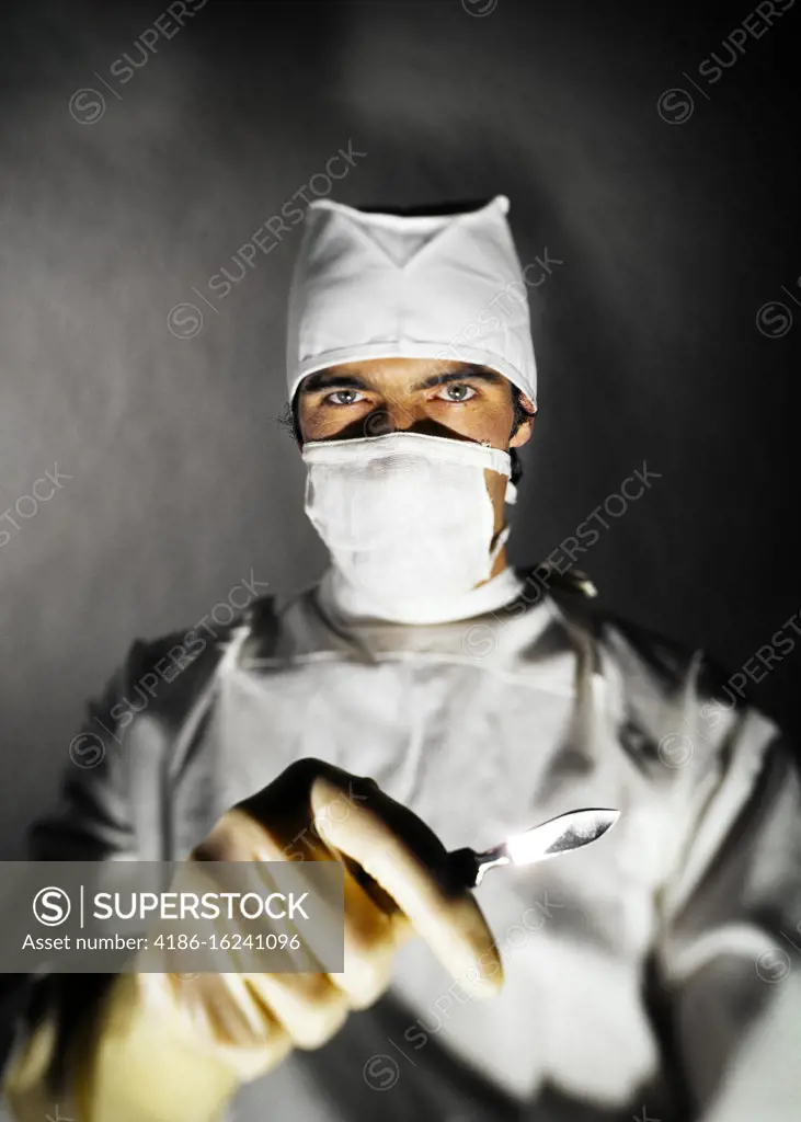 1970s INTENSE MAN DOCTOR SURGEON HOLDING SCALPEL IN GLOVED HAND WEARING SCRUBS AND MASK STARING DIRECTLY LOOKING AT CAMERA
