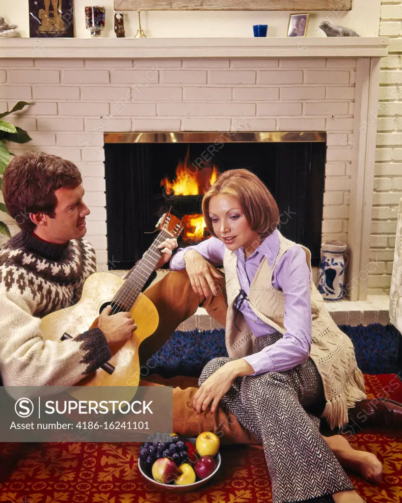 1960s 1970s COUPLE SITTING TOGETHER BY BRICK FIREPLACE MAN PLAYING GUITAR BAREFOOT WOMAN WEARING BELLBOTTOMS LISTENING ENTRANCED