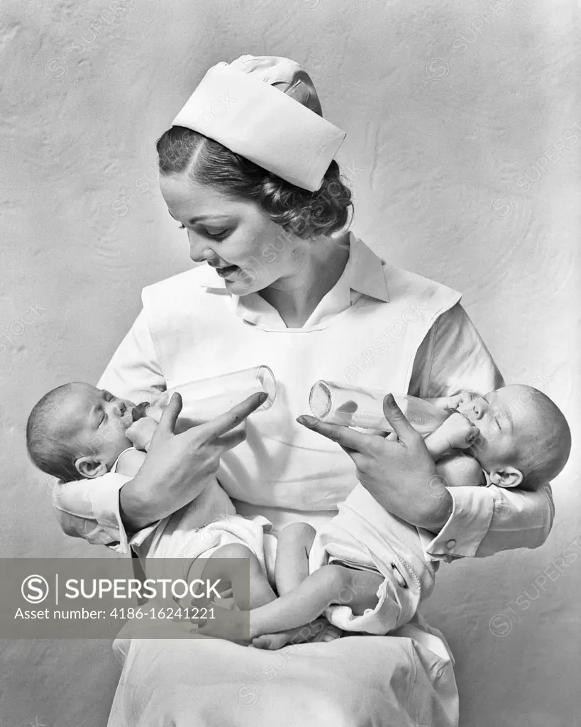 1930s SMILING NURSE SITTING HOLDING JUGGLING TWO NEWBORN TWIN BABY GIRL SISTERS IN HER ARMS FEEDING THEM WITH BOTTLES
