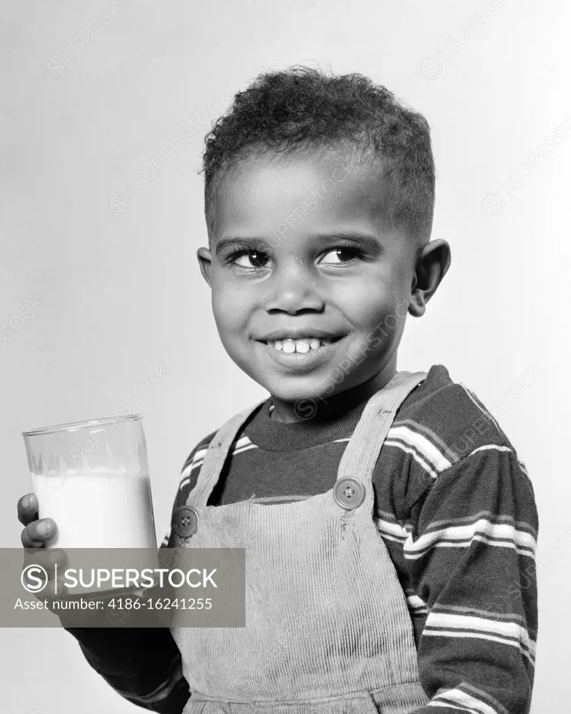 1940s 1950s SMILING AFRICAN-AMERICAN BOY HOLDING DRINKING GLASS OF MILK WEARING BIB OVERALL STRIPED T-SHIRT LOOKING OFF TO SIDE