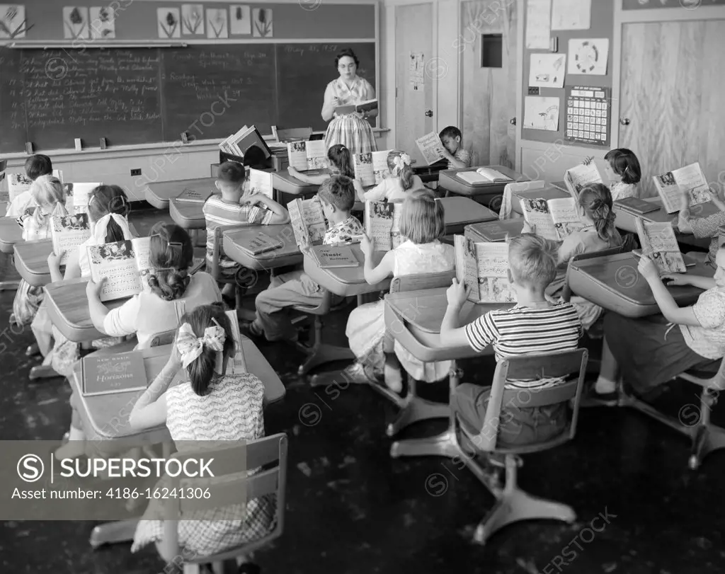 1950s 1960s ELEMENTARY SCHOOL CLASSROOM TEACHER WOMAN AND STUDENTS READING FROM OPEN TEXTBOOKS LEARNING BLACKBOARD DESKS