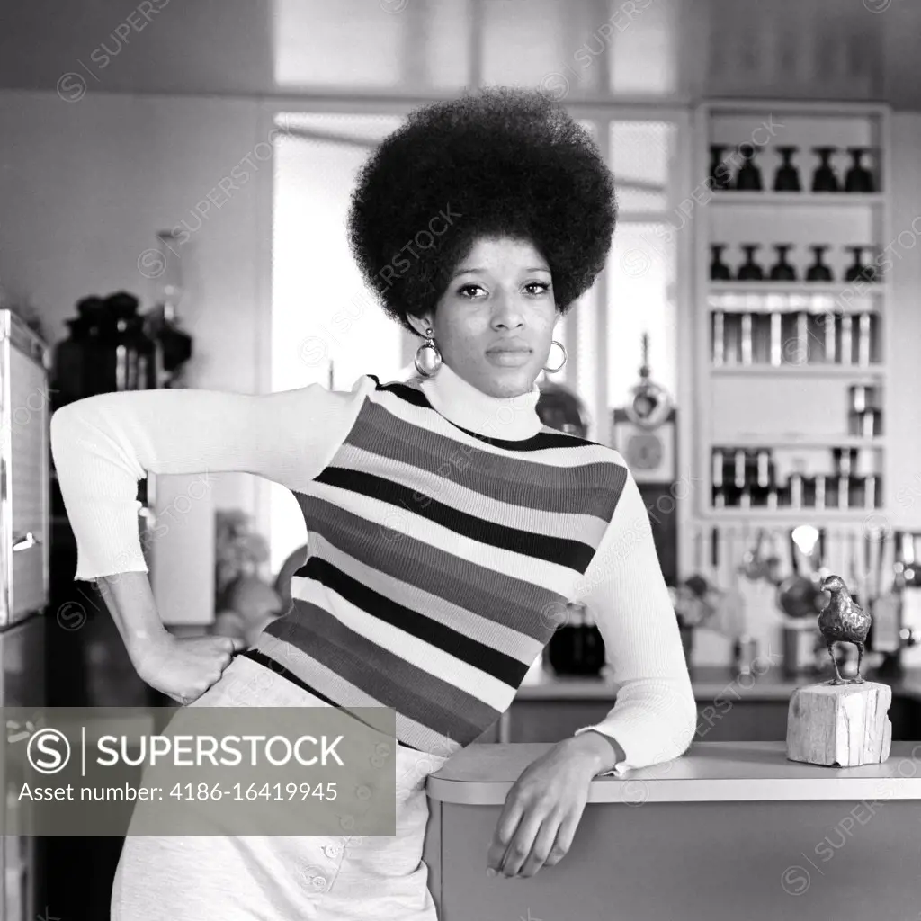1960s 1970s AFRICAN-AMERICAN WOMAN TALL AFRO HAIRSTYLE STRIPED SHIRT LEANING ON RESTAURANT COUNTER HAND ON HIP LOOKING AT CAMERA