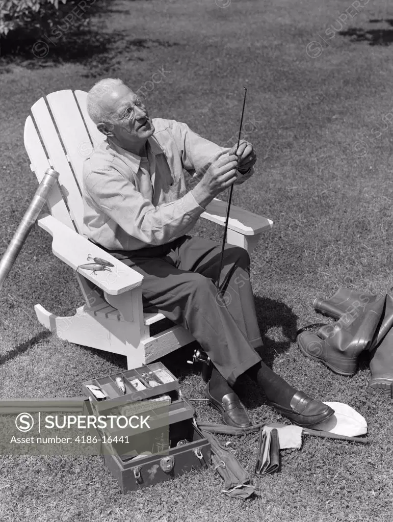 1950S Elderly Man In Backyard Seated In Wooden Chair Threading Line Through Fishing Rod With Boots & Tackle Box At Feet