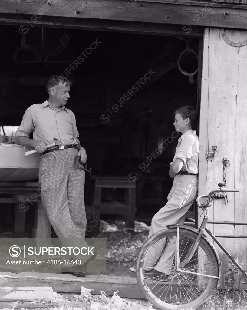1930S Man Father Holding Hand Tools Talking To Boy Son Leaning In Doorway Of Boat Shed