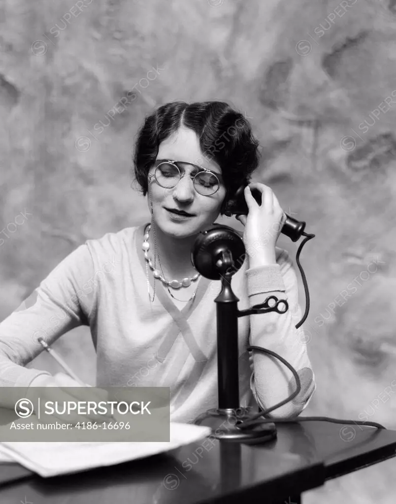 1920S Woman Wearing Pince-Nez Sitting At Desk Talking On Candlestick Phone & Writing