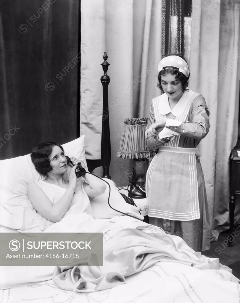 1920S 1930S Two Women In Bedroom Maid Looking At Wrist Watch Other Woman In Bed Talking On Phone Hand Held Over Mouthpiece
