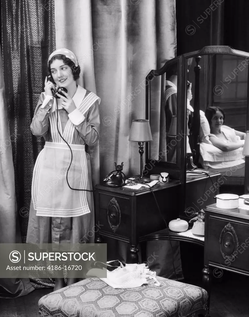 1920S 1930S Maid In Uniform Talks On Telephone In Front Of Vanity Dressing Table Other Woman Is Seen As Reflection In Mirror