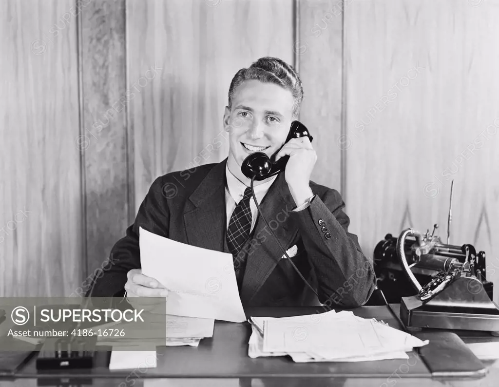 1930S 1940S Smiling Businessman Talking On Telephone Sitting At Desk With Dictation Machine 