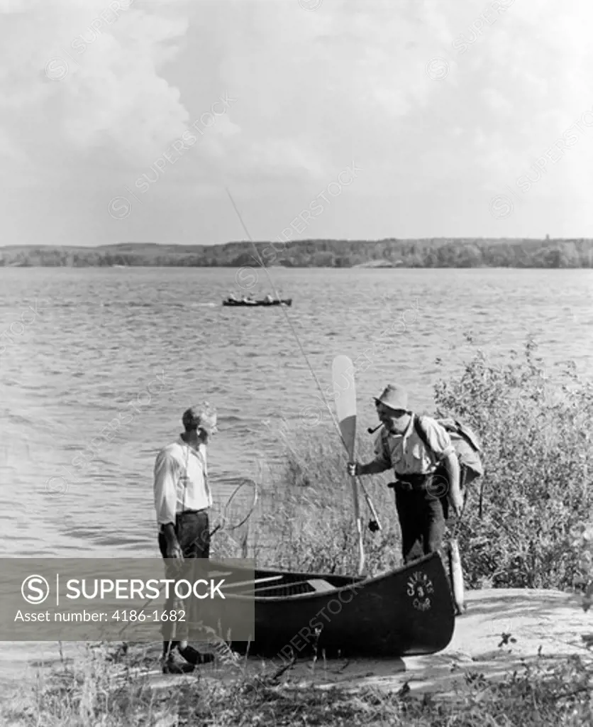 1940S 1950S Two Men Standing By River Next To Canoe One Man Holding Oar Lake Of The Woods Ontario Canada