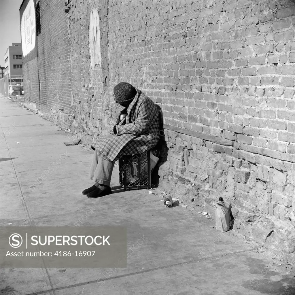 1970S 1980S Homeless Person Bundled In Winter Coat & Hat Sitting On Milk Crate Against Brick Wall With Head Bowed Down