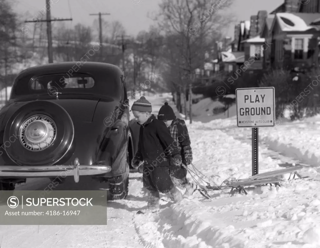 1940S Two Boys Pulling Sleds Across A Snowy Suburban Winter Street With Parked Car And Sign That Says Play Ground