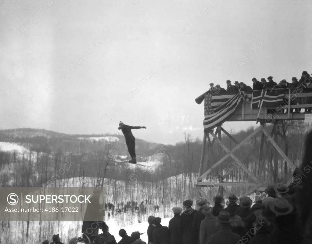 1930S Man Ski Jumping In Mid Air Crowd Spectators Grand Stand Draped With Flags And Bunting Winter Snowy Landscape