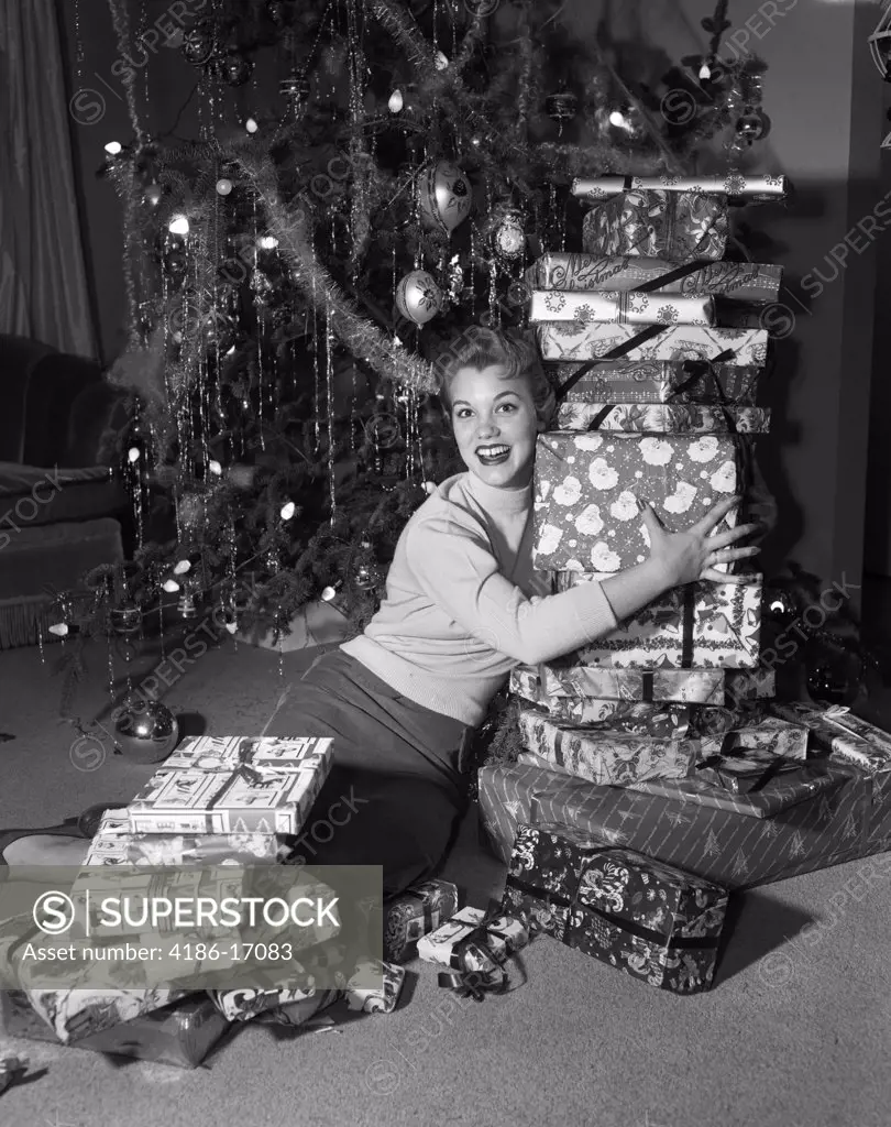 1950S Young Smiling Woman Sitting On Floor By Christmas Tree Holding Up Tall Stack Of Presents Looking At Camera
