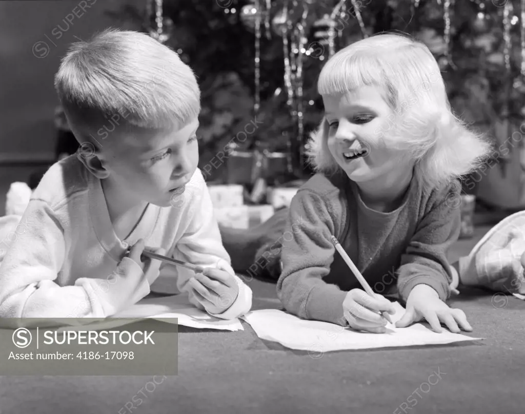 1960S Boy And Smiling Girl Laying On Floor Each With A Pencil And Sheet Of Paper Writing Christmas Tree In Background