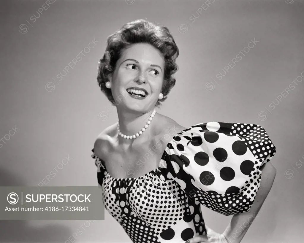 1950s SMILING WOMAN LOOKING OVER HER SHOULDER POSING WEARING LOW CUT SCOOP NECKLINE DRESS WITH POLKA DOTS AND PUFFY SLEEVES