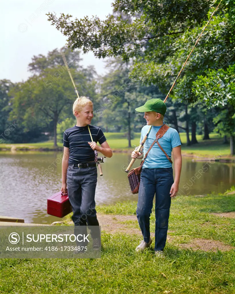 1970s TWO BOYS WALKING ALONG LAKESIDE TALKING CARRYING FISHING POLES AND TACKLE BOXES