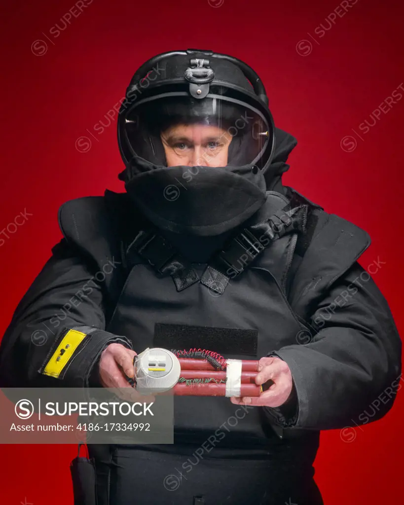 1990s MAN BOMB SQUAD TECHNICIAN LOOKING AT THE CAMERA HOLDING A BOMB STICKS OF EXPLOSIVE WITH WIRES AND TIMER ATTACHED