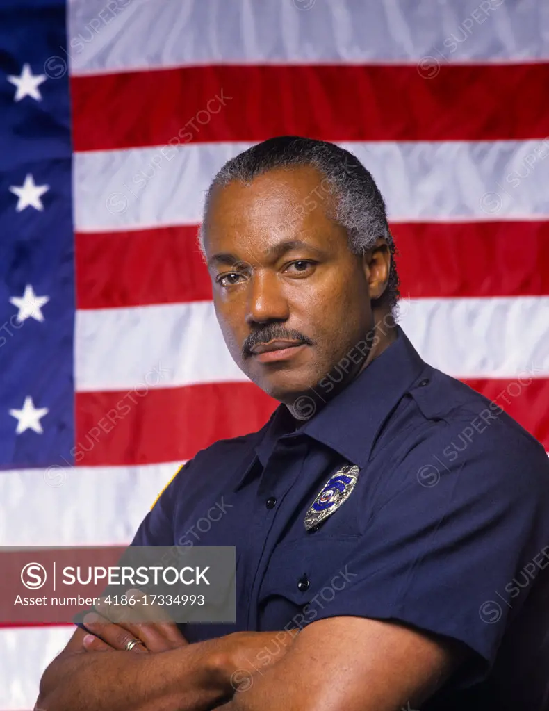 1990s BLACK POLICEMAN LOOKING AT CAMERA STANDING IN FRONT OF U.S. FLAG 