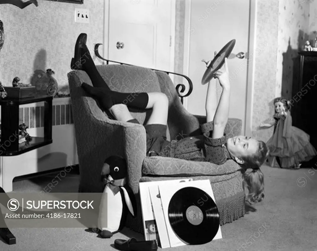 1950s 1960s TEENAGE GIRL SMILING RECLINING UPSIDE DOWN ON CHAIR HOLDING VINYL RECORD ALBUM LISTENING TO MUSIC