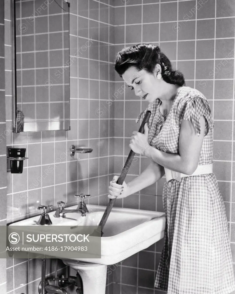 1940s ANNOYED EXASPERATED BRUNETTE WOMAN HOUSEWIFE IN THE BATHROOM USING A HAND PLUNGER TO CLEAR A CLOGGED SINK DRAIN