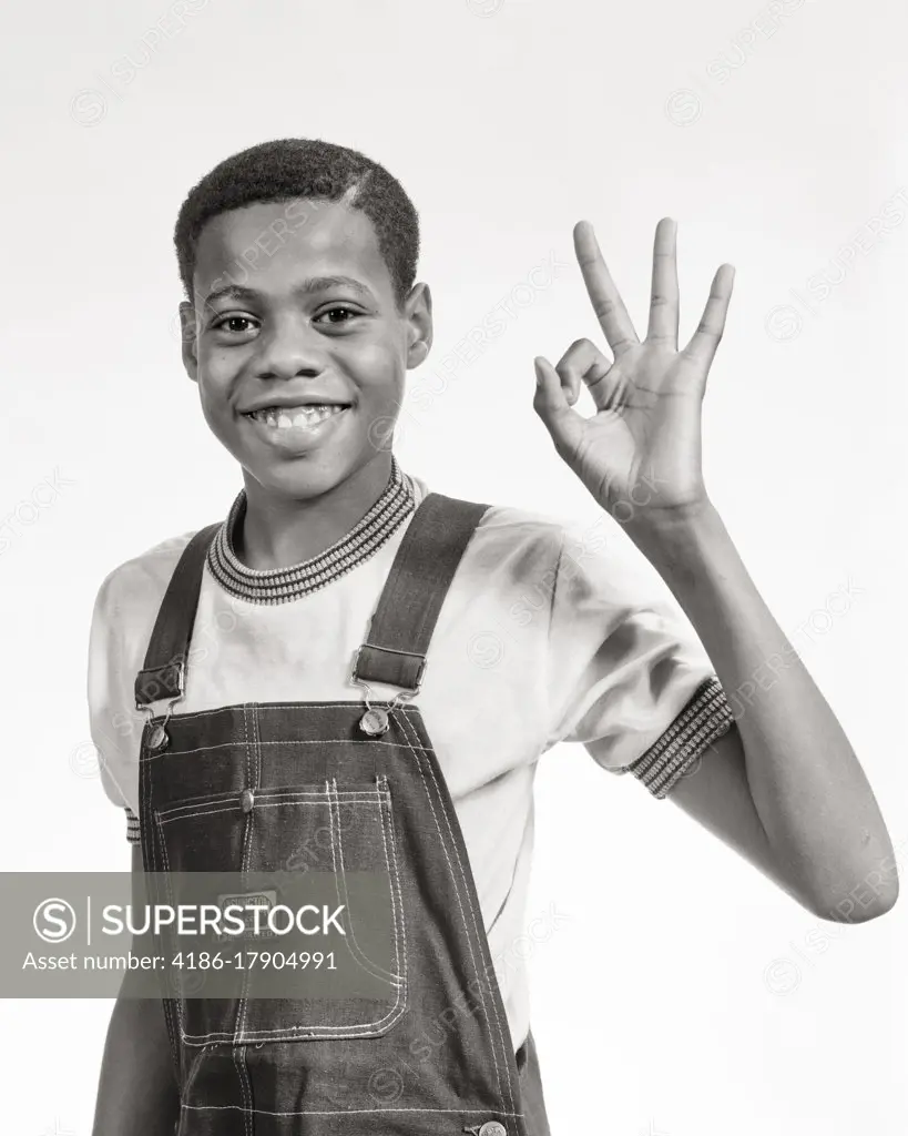 1970s SMILING AFRICAN-AMERICAN PRE-TEEN BOY WEARING BIB OVERALLS MAKING OKAY SIGN WITH HAND LOOKING AT CAMERA
