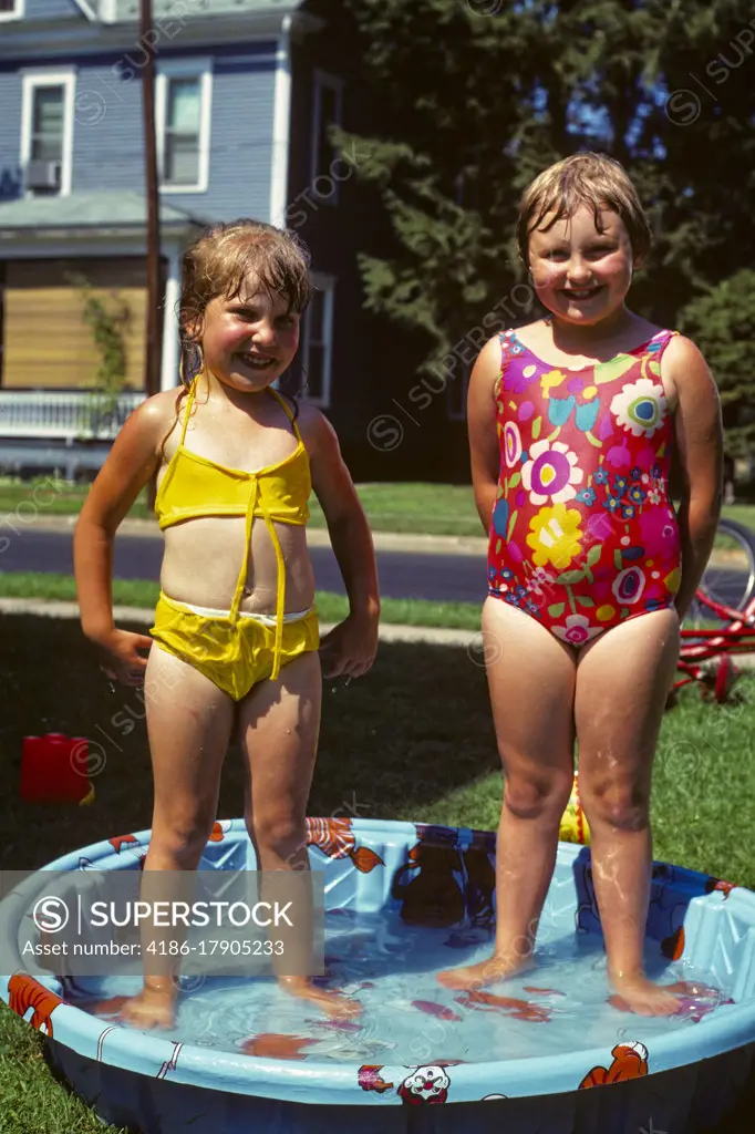 1970s TWO SMILING GIRLS WEARING MESSY WET BATHING SUITS YELLOW AND RED STANDING IN KIDDIE POOL IN BACKYARD LOOKING AT CAMERA 
