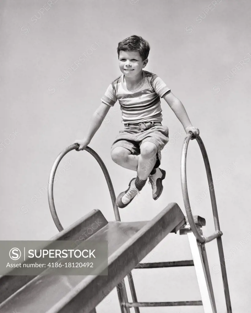 1940s BOY AT TOP OF PLAYGROUND SLIDE PUSHING UP ON HAND RAILS CROSSING HIS LEGS FUNNY FACIAL EXPRESSION 