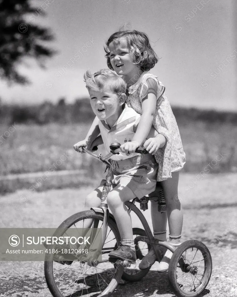 1930s BOY AND GIRL BROTHER SISTER RIDING TANDEM ON TRICYCLE GIRL STEERING ON BACK OF TRIKE BOY PEDDLING LAUGHING HAVING FUN 