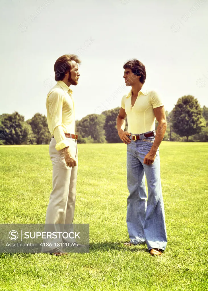 1970s TWO YOUNG MEN WEARING BELL BOTTOM PANTS STANDING ON GRASSY HILL TALKING TOGETHER