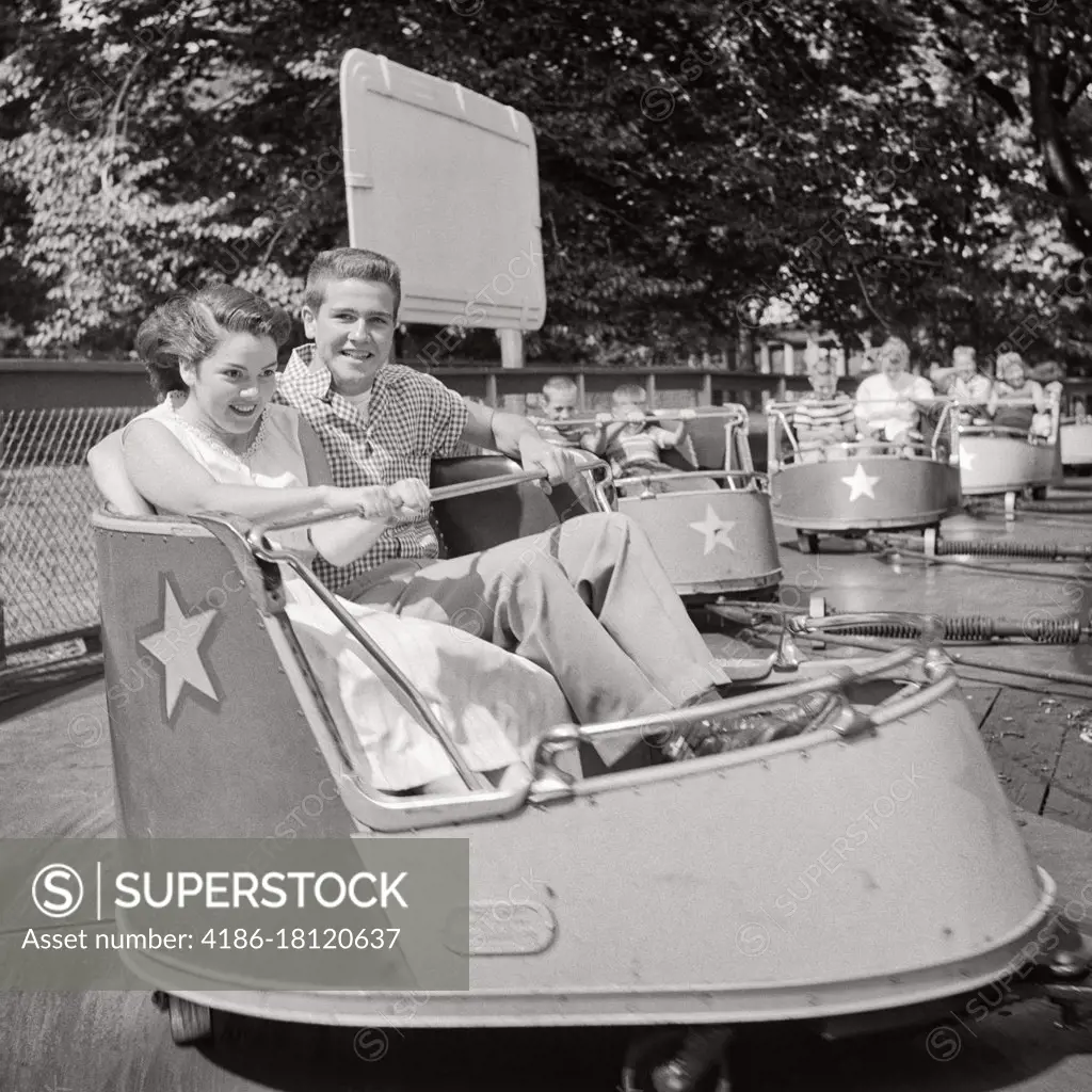 1950s TEEN COUPLE AT AMUSEMENT PARK RIDING IN THE WHIP LOOKING AT CAMERA
