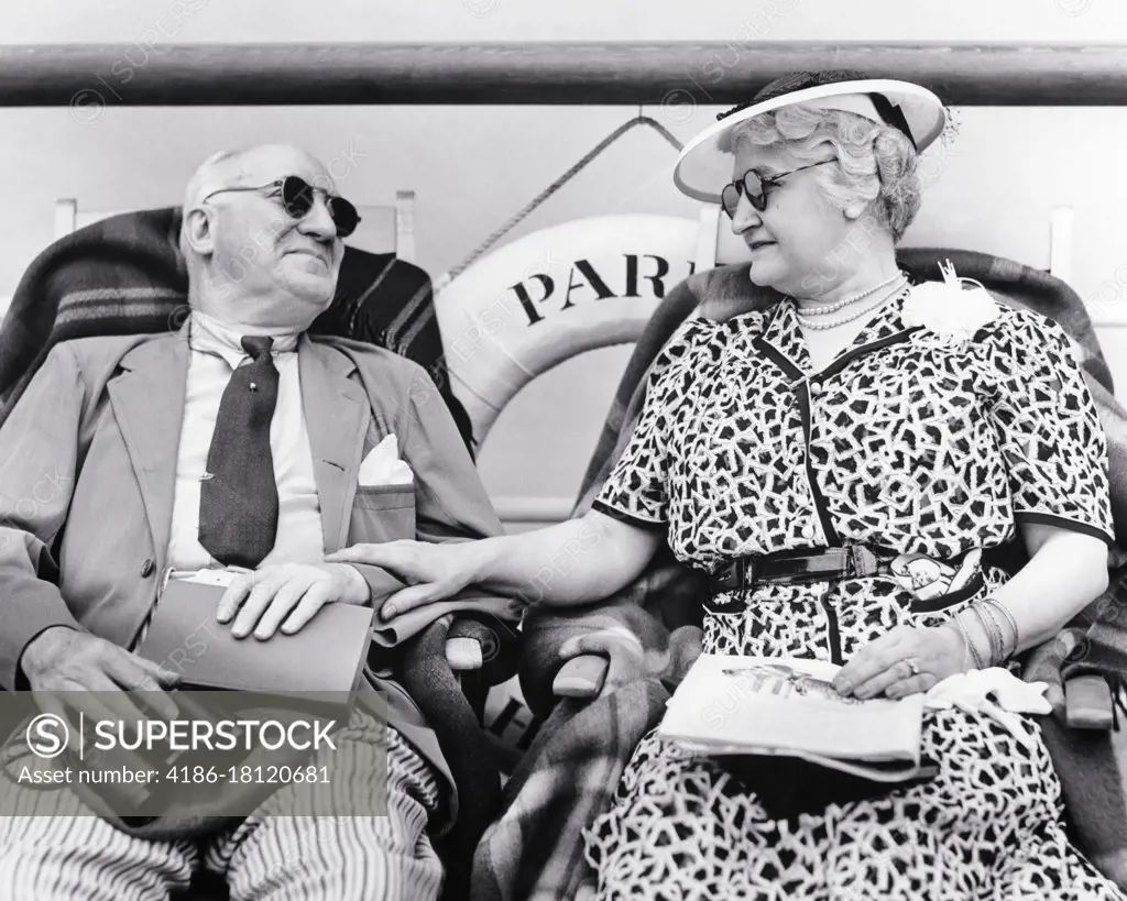 1930s SENIOR RETIRED COUPLE FASHIONABLY DRESSED SITTING IN DECK CHAIRS SIDE BY SIDE ON A CRUISE SHIP BOTH WEARING SUNGLASSES