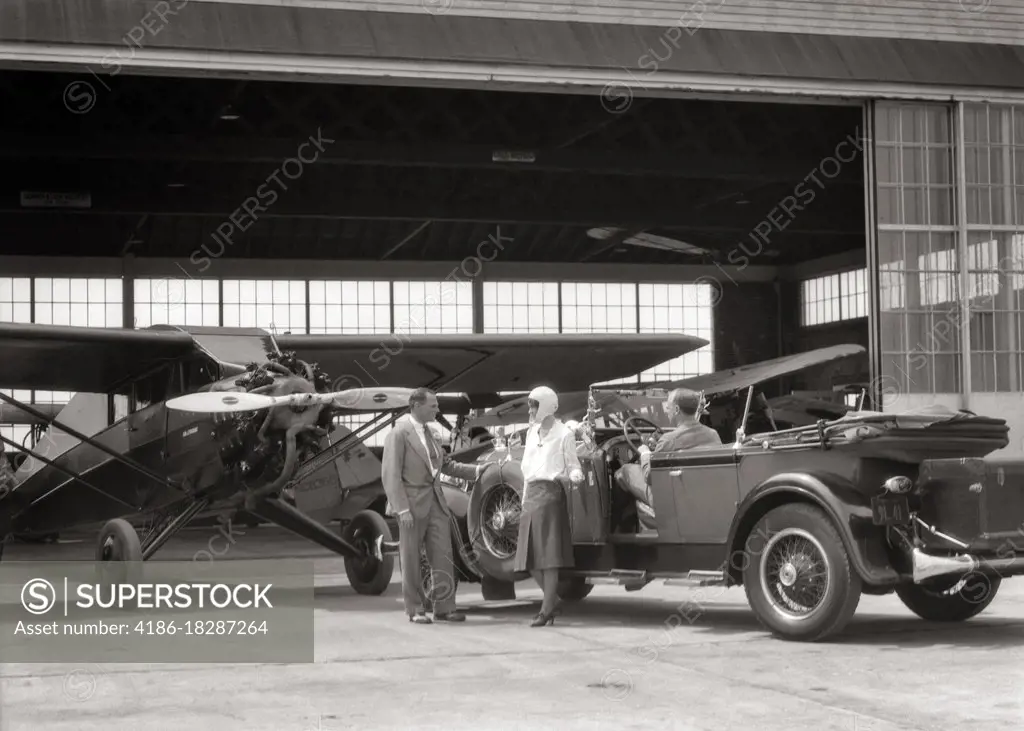1920s 1930s COUPLE WITH CONVERTIBLE PACKARD CAR TALKING TO AIRPLANE SALESMAN AT HANGAR 