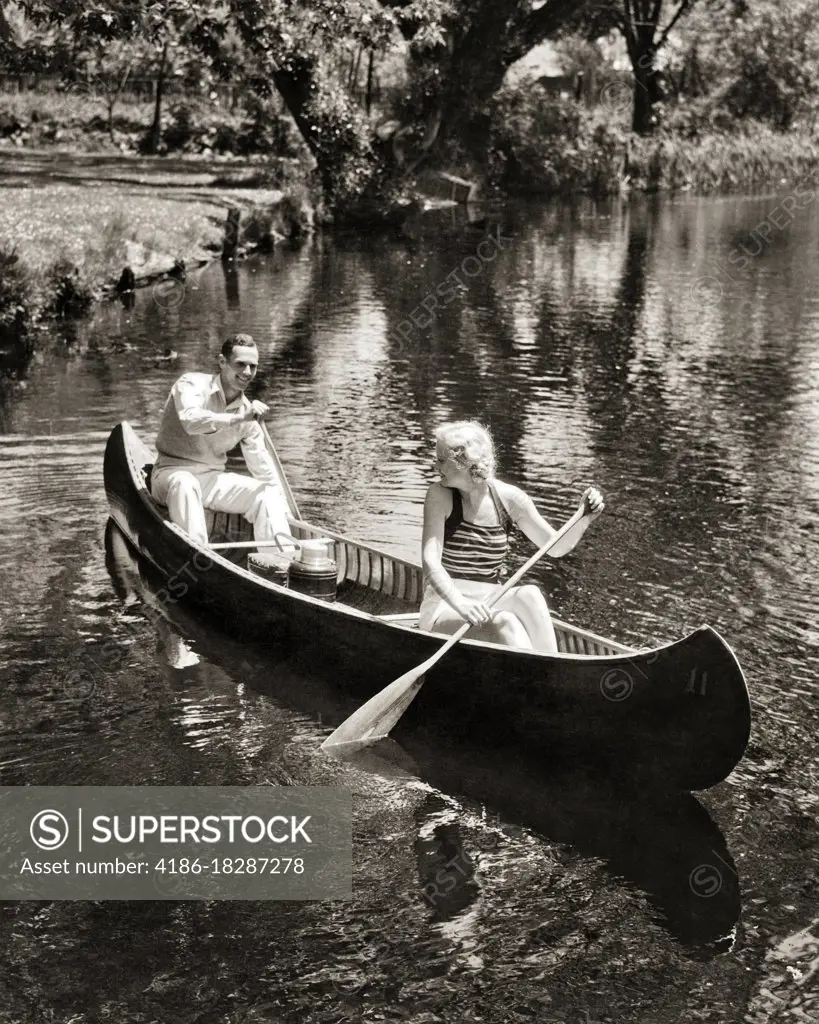 1930s COUPLE MAN AND WOMAN PADDLING A CANOE GOING ON A SUMMER PICNIC WITH BASKET AND THERMOS 
