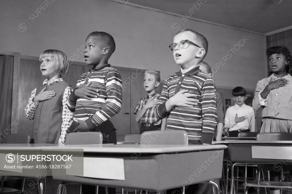 1960s GRADE SCHOOL KIDS STANDING AT DESKS SAYING PLEDGE OF ALLEGIANCE TO UNITED STATES FLAG WITH HANDS OVER THEIR HEARTS