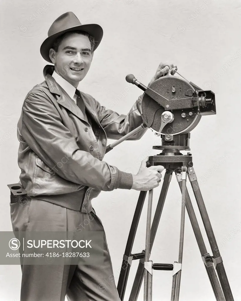 1930s SMILING MAN CINEMATOGRAPHER OPERATING AKELEY HAND CRANK MOVIE CAMERA LOOKING AT CAMERA WEARING LEATHER JACKET FEDORA HAT