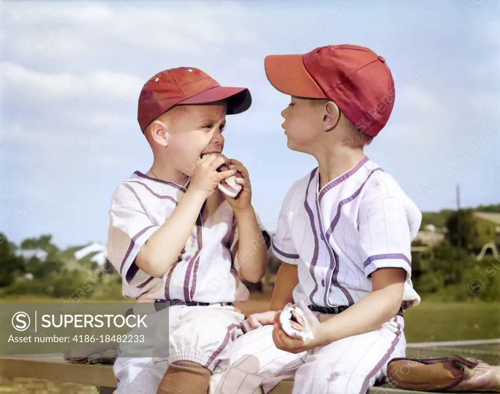 1960s TWO BOYS IN LITTLE LEAGUE BASEBALL CAPS AND UNIFORMS SITTING TOGETHER EATING HOT DOGS