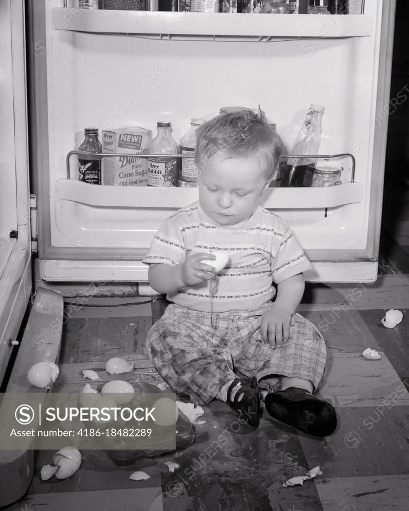 1950s INQUISITIVE MESSY BOY TODDLER ON KITCHEN FLOOR IN FRONT OF AN OPEN REFRIGERATOR DOOR PLAYING WITH TRAY OF BROKEN EGGS 