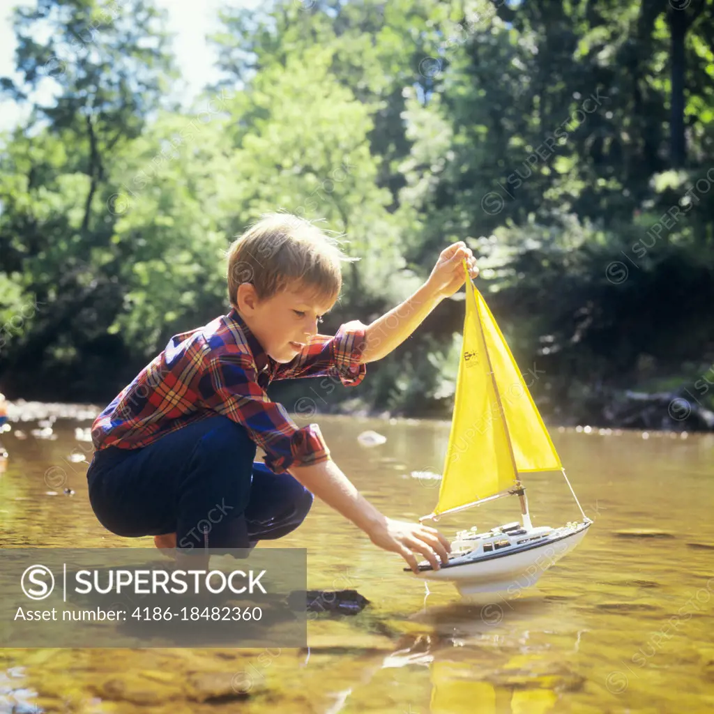 1970s BOY ABOUT TO LAUNCH A TOY SAIL BOAT INTO A STREAM