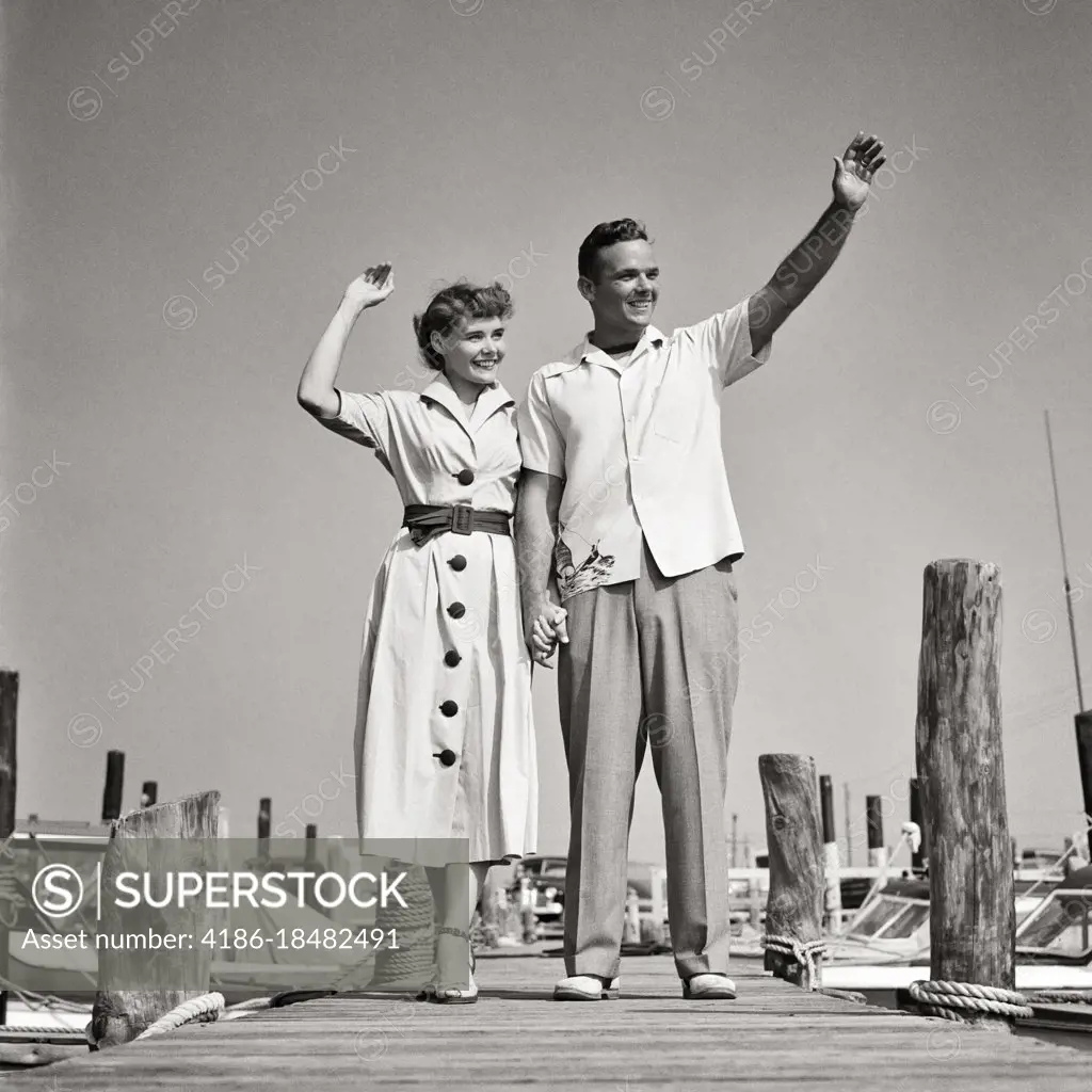1940s 1950s YOUNG COUPLE WEARING STYLISH SUMMER CLOTHES STANDING ON WOODEN DOCK WAVING HELLO GOODBYE GOOD-BYE 