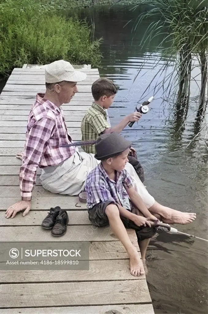1950s 1960s FATHER WITH TWO SONS SITTING ON DOCK FISHING TOGETHER OUTDOOR BY POND