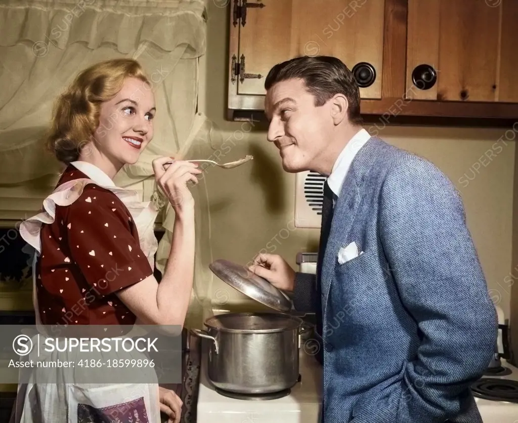 1950s SMILING HOUSEWIFE AT STOVE GIVING HAPPY HUSBAND TASTE OF HER COOKING