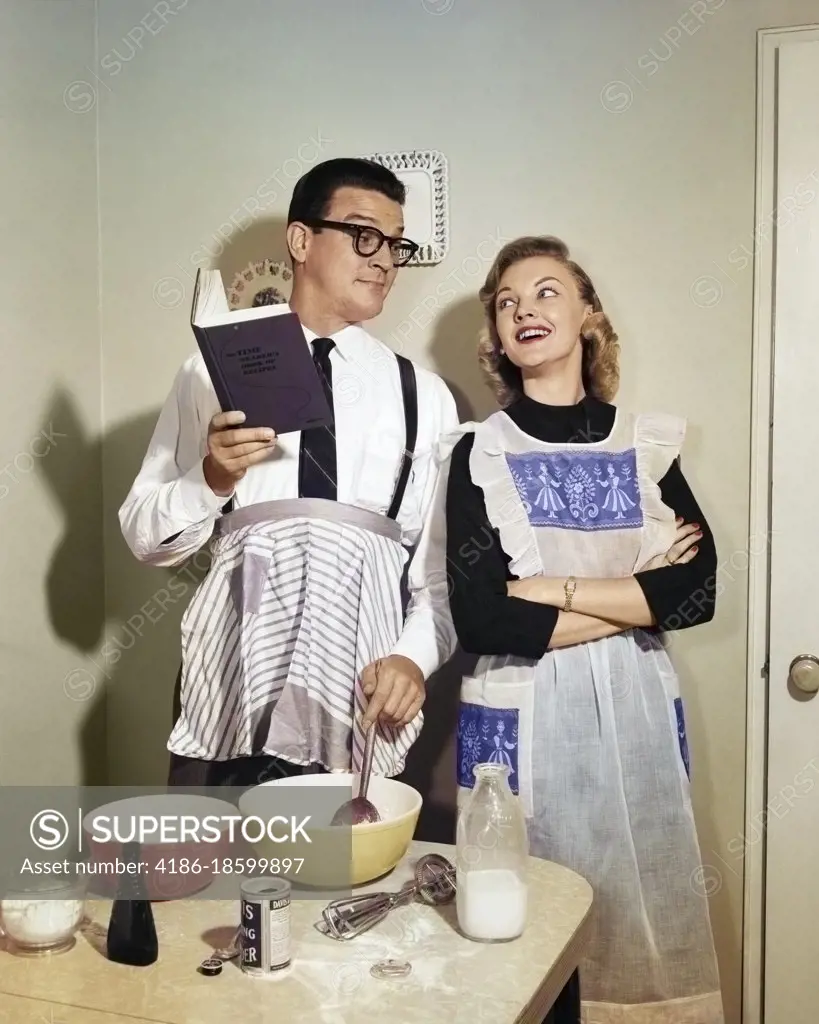 1950s SMILING COUPLE IN KITCHEN WITH HUSBAND LEARNING TO COOK FROM A BOOK WHILE WIFE WATCHES BOTH WEARING APRONS