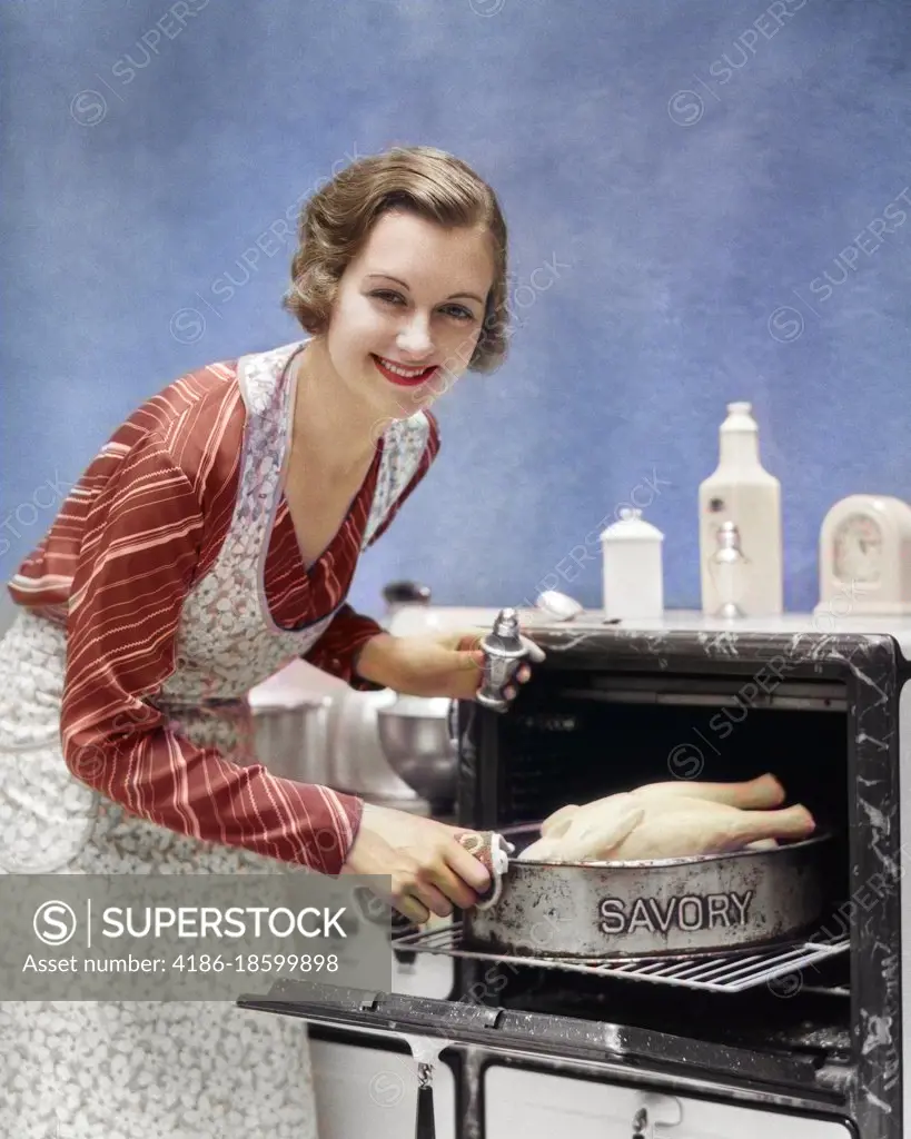 1930s SMILING HOUSEWIFE IN APRON LOOKING AT CAMERA SALTING SEASONING TURKEY IN SAVORY ROASTING PAN GOING INTO OVEN