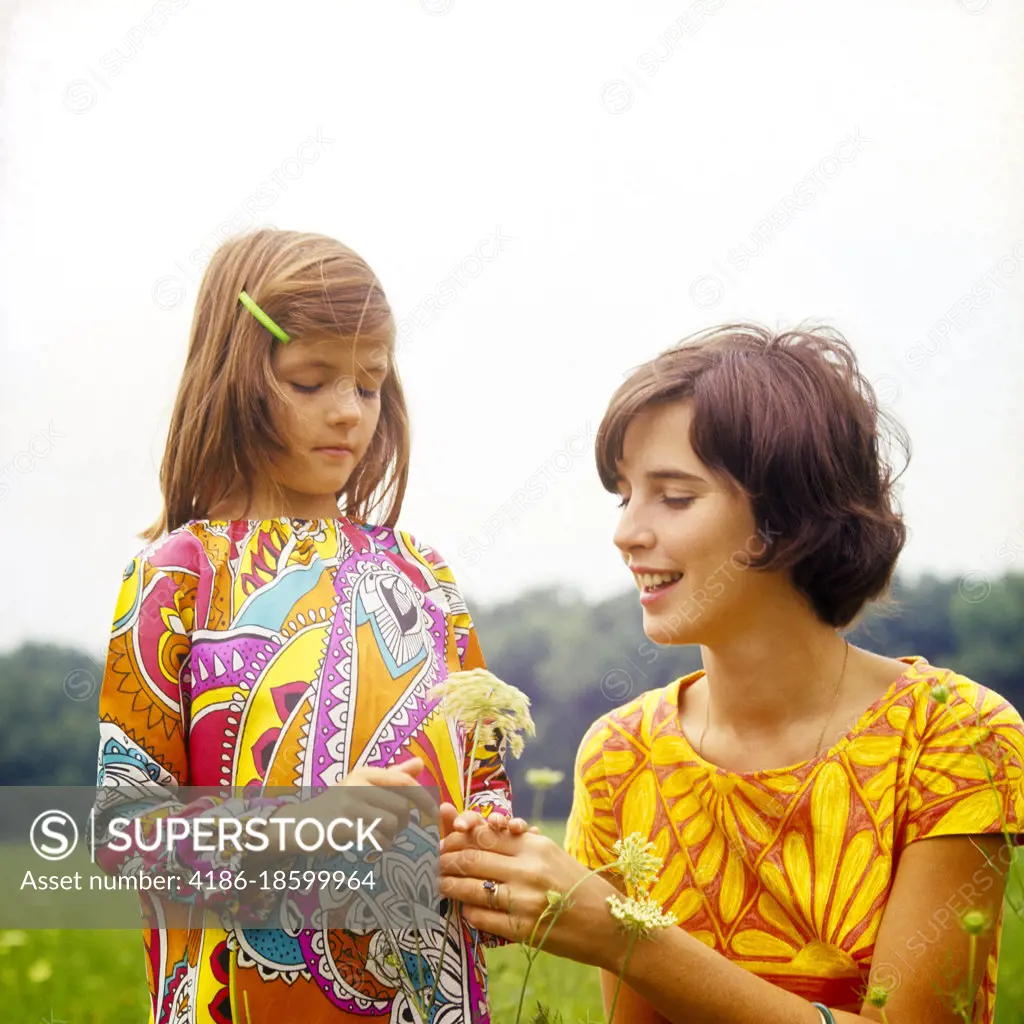 1960s 1970s BRUNETTE MOTHER AND CURIOUS DAUGHTER WEARING PRINT DRESSES OUTDOOR EXAMINING PICKING QUEEN ANNES LACE WILD FLOWER
