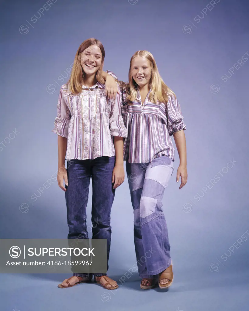 1970s TWO TEENAGE GIRLS STANDING LEANING ON EACH OTHER SMILING WEARING BELL-BOTTOM DENIM AND STRIPED SHIRTS LOOKING AT CAMERA