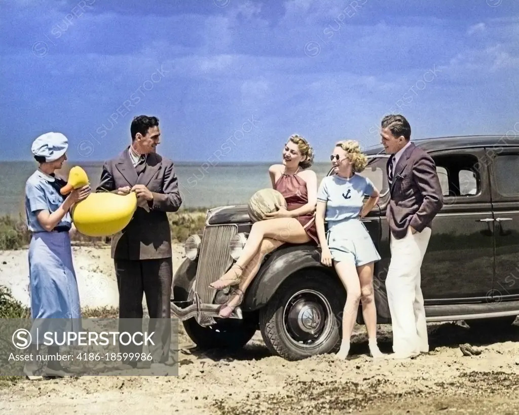 1930s FIVE MEN & WOMEN IN CASUAL SUMMER CLOTHES PLAYING WITH INFLATABLE BEACH TOYS BY 1936 FORD V-8 SEDAN PARKED ALONG SEA SHORE