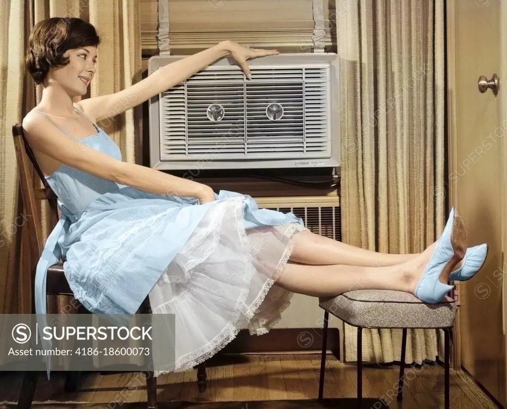 1960s SMILING WOMAN SITTING PROPPING LEGS UP ON STOOL RESTING HAND ON TOP OF LOOKING AT AIR CONDITIONER
