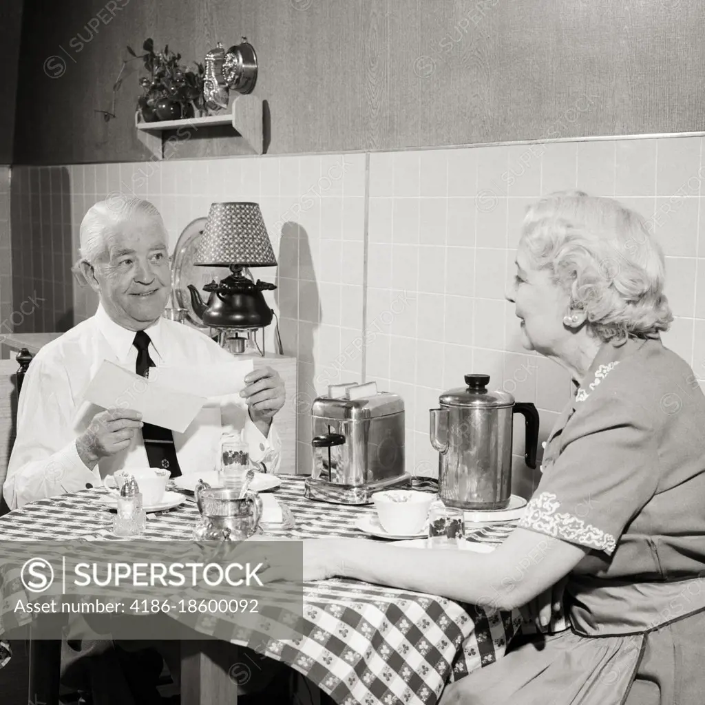 1950s SENIOR COUPLE SITTING AT KITCHEN TABLE HAVING TOAST AND COFFEE MAN OPENING MORNING MAIL SMILING HOLDING CHECK