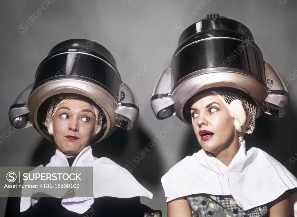 1950s TWO WOMEN GOSSIPING SITTING TOGETHER UNDER HAIR DRYERS IN HAIRDRESSER SALON